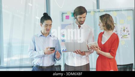 Presenting woman in red dress with team discussing laptop tablet phone in office near whiteboard Stock Photo
