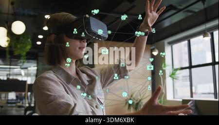 Gesturing woman wearing beige shirt manipulating holographic icons in modern office with VR headset Stock Photo