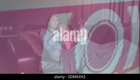 Watching two women wearing denim on leather sofa at home, viewing projection, holding popcorn tub Stock Photo