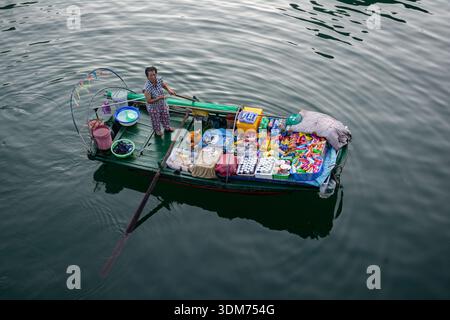 A lone vendor stands on a green wooden boat, selling a colorful array of snacks, drinks, and small goods at a floating market. Calm water creates a tr Stock Photo