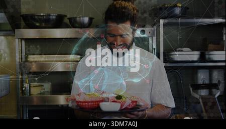 Holding bearded chef in white coat presenting tray at kitchen, with checked baskets and hologram Stock Photo