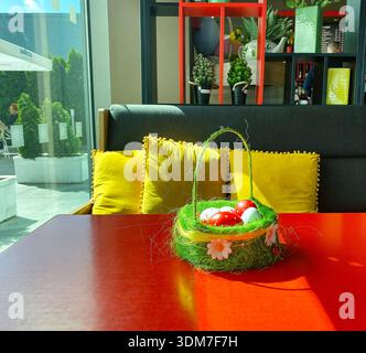 Colorful Easter basket filled with decorated eggs placed on a red table in a modern cafe, featuring bright cushions and greenery nearby Stock Photo