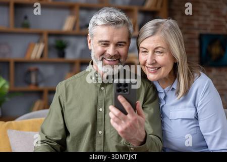 Happy senior couple sharing a moment, smiling and connecting with loved ones through a modern smartphone video call at home, embracing technology for communication and family bond Stock Photo