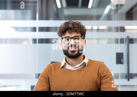 Confident young man with curly hair and beard wearing glasses, smiling directly at the viewer, showing a positive and approachable presence in a contemporary workplace environment Stock Photo