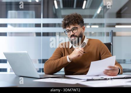 Young businessman in smart-casual attire studies reports on a laptop, holding a pen and papers while analyzing financial data and planning strategy in a modern office setting Stock Photo