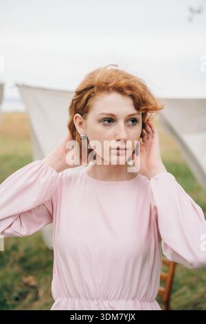 Natural portrait of a young red-haired woman adjusting her hair while standing outdoors. She wears a soft pink dress and minimal jewelry, surrounded b Stock Photo