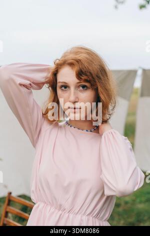 A red-haired woman in a light pink dress poses outdoors wearing a handmade blue beaded necklace. The natural lighting and minimal background highlight Stock Photo