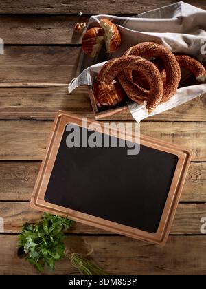 Fresh bakery simit with empty serving plate and herbs on rustic wooden table – bakery food styling Stock Photo
