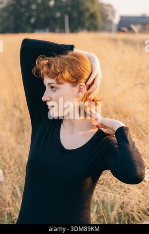 Portrait of a young red-haired woman wearing elegant gold earrings. She poses with arms raised in a sunlit field, highlighting the accessory. The soft Stock Photo