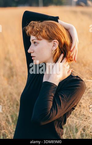 Portrait of a young red-haired woman wearing elegant gold earrings. She poses with arms raised in a sunlit field, highlighting the accessory. The soft Stock Photo