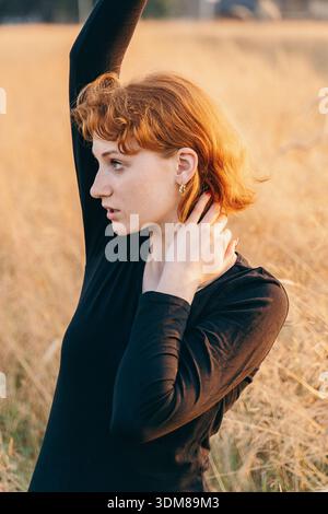 Portrait of a young red-haired woman wearing elegant gold earrings. She poses with arms raised in a sunlit field, highlighting the accessory. The soft Stock Photo