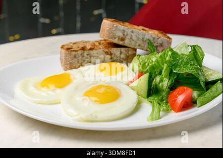 Breakfast of scrambled eggs with tomato and cucumber salad on toasted whole grain bread. A plate of fried eggs, vegetable salad and crispy baguette in Stock Photo