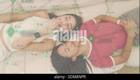 Lying two women talking on sofa cushions, wearing smartwatches, crop top, red tee, green overlay Stock Photo