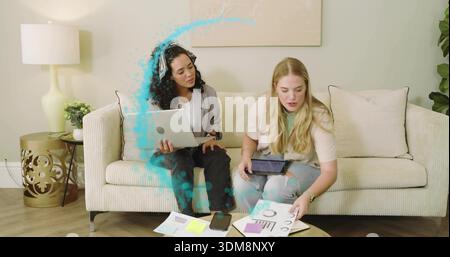 Collaborating, two women in blazer and jeans reviewing charts on beige sofa, with laptop and tablet Stock Photo