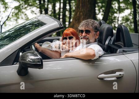 Wearing sunglasses, side view. Senior couple driving convertible cabriolet car. Stock Photo