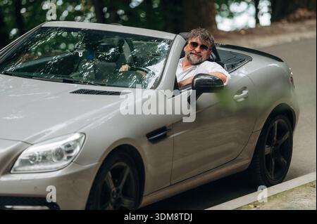 Looking at the camera. Stylish senior man is in convertible cabriolet car. Stock Photo