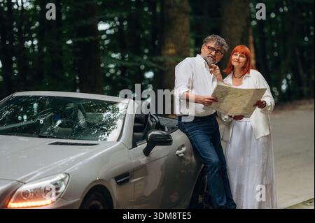 Reading the map. Lovely senior couple standing on the road near convertible cabriolet car. Stock Photo