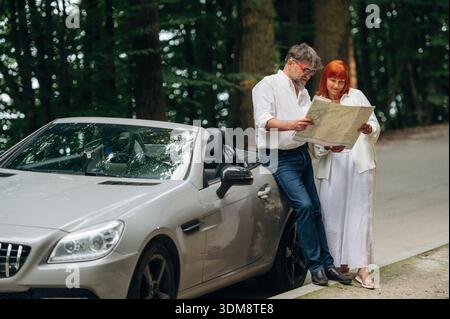 Reading the map. Lovely senior couple standing on the road near convertible cabriolet car. Stock Photo
