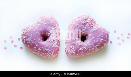 Two heart-shaped donuts with pink icing and sprinkles, isolated on a white background with pink confetti. Romantic or Valentine's Day theme. Stock Photo
