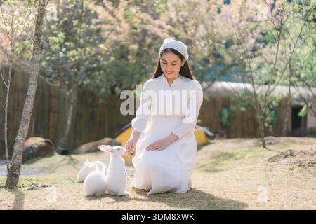Happy beautiful woman in white dress feeding cute white rabbits in green garden captured in soft natural light, representing kindness, lifestyle, and Stock Photo