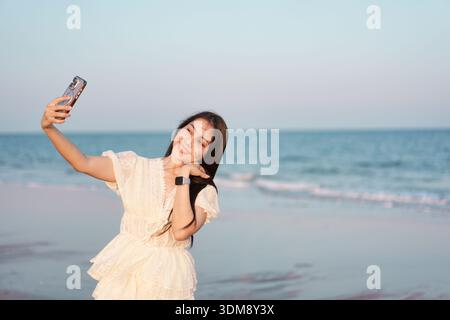 Beautiful asian woman in white dress enjoying while taking selfie with smartphone on the seaside beach. creating joyful travel and leisure atmosphere Stock Photo