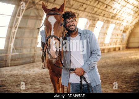 Front view, standing together. Man is with horse in the hangar. Stock Photo