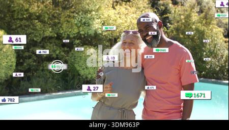 Smiling senior couple standing poolside in yard wearing pink tee, grey dress, shades, social badges Stock Photo