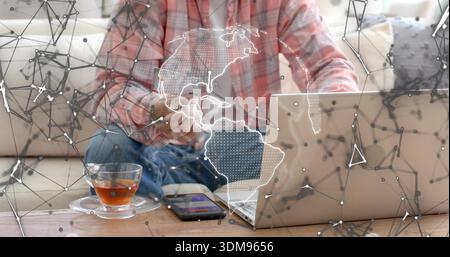 Using laptop, holding card, Indian man wearing plaid shirt on sofa with phone, teacup, map overlay Stock Photo