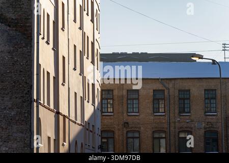 Sunlit city architecture with beige apartment wall casting shadows onto brick industrial building, tall windows, street light and overhead wires Stock Photo