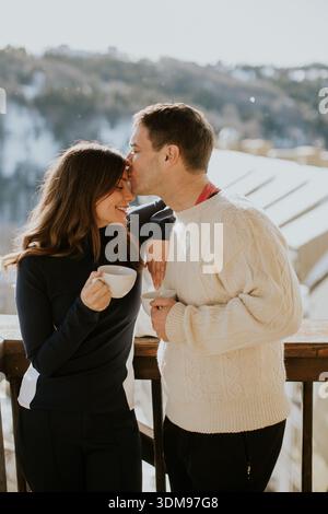 A joyful couple shares warm drinks while enjoying the beautiful snowy landscape outside. Their smiles shine brighter than the sun. Stock Photo