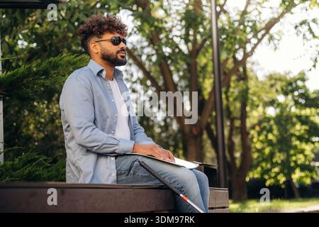 Sitting and reading using Braille. Blind man with white cane and sunglasses is in the city. Stock Photo
