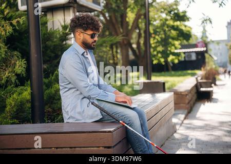 Sitting and reading using Braille. Blind man with white cane and sunglasses is in the city. Stock Photo