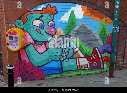 Hammo street art mural of a Troll cartoon figure using a mobile phone beside a wall-mounted public defibrillator (AED) on arch Stockport Stock Photo