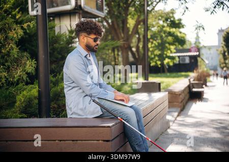 Sitting and reading using Braille. Blind man with white cane and sunglasses is in the city. Stock Photo