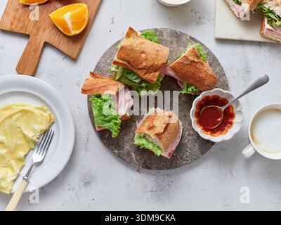 Freshly made sandwiches with ham, mozzarella, and lettuce are arranged on a table with sliced oranges. Tomato sauce and butter are also served alongsi Stock Photo