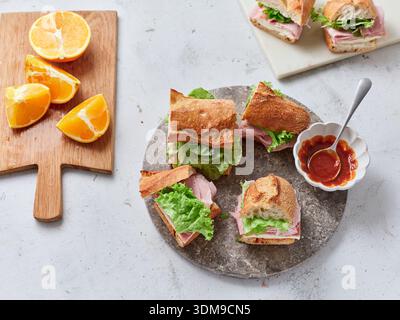 Sliced baguette sandwiches are arranged on a stone plate. Fresh oranges are next to the sandwiches. A small bowl of sauce is also present on the table Stock Photo