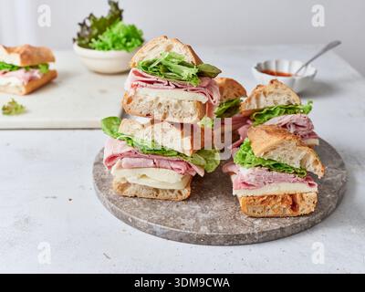 Freshly made sandwiches stacked on a stone platter. Ingredients include baguette, mozzarella cheese, ham, lettuce, and tomato sauce. Stock Photo