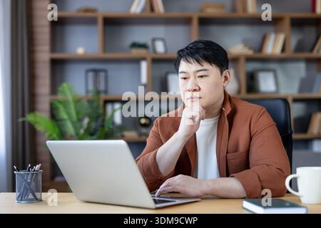 Asian man sitting at a desk, looking at his laptop screen with a pensive expression, representing concentration, remote work, and focused digital activity from a comfortable home office setting Stock Photo