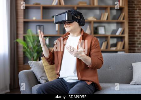 Man wearing a virtual reality headset and headphones, actively interacting with a digital world from his living room couch, enjoying immersive entertainment and advanced technology Stock Photo