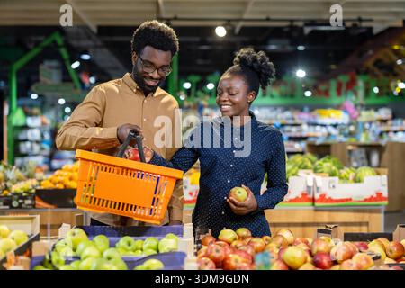 Young couple picking fresh red and green apples from a display in a bright supermarket produce department, enjoying a healthy lifestyle and shopping for groceries together Stock Photo
