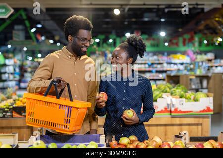 Young couple smiling, enjoying grocery shopping together, selecting fresh apples and other healthy fruits in the produce section of a modern supermarket, focusing on a healthy lifestyle Stock Photo