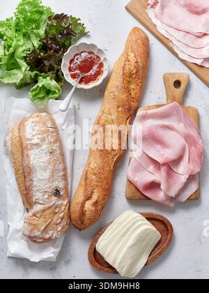 A lunch or breakfast table contains sliced baguette, ham, mozzarella, lettuce, tomato sauce for preparation and serving. Stock Photo