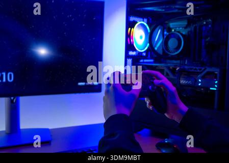 Gamer holding game controller while playing video game on computer monitor next to illuminated gaming pc in dark modern room Stock Photo