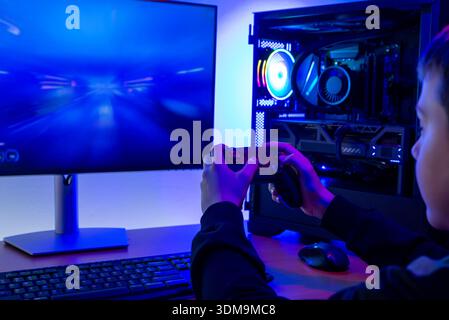 Gamer holding game controller while playing video game on computer monitor next to illuminated gaming pc in home entertainment environment Stock Photo