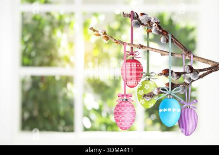 Easter eggs hanging from willow branch near window indoors Stock Photo