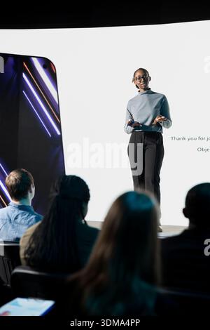 Black female product manager presenting new smartphone reveal with detailed presentation about interface and other modern features. Launch event at convention with audience group. Stock Photo