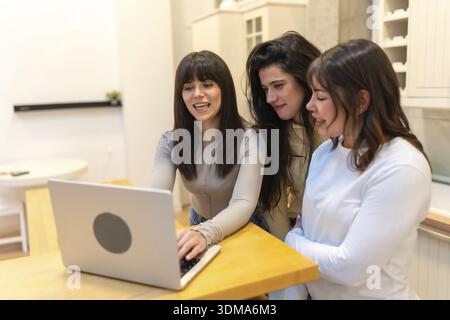 Three friends, women, collaborating and smiling while looking at a laptop computer on a wooden counter, sharing a moment of remote work or online conn Stock Photo