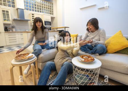 Three young women lounging on a sofa and floor, sharing pizza, laughing and chatting in a cozy apartment, enjoying relaxed weekend togetherness and fr Stock Photo