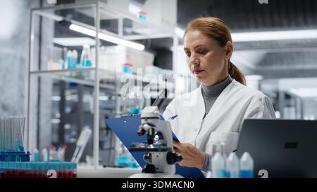 Hematology specialist in lab conducting research on blood sample, taking notes. Epidemiologist in laboratory analyzing sanguine fluid specimen in vial, writing down findings, camera B Stock Photo