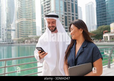 Arab businessman showing smartphone content to an Asian businesswoman as they walk through the urban landscape of Dubai Marina. Stock Photo